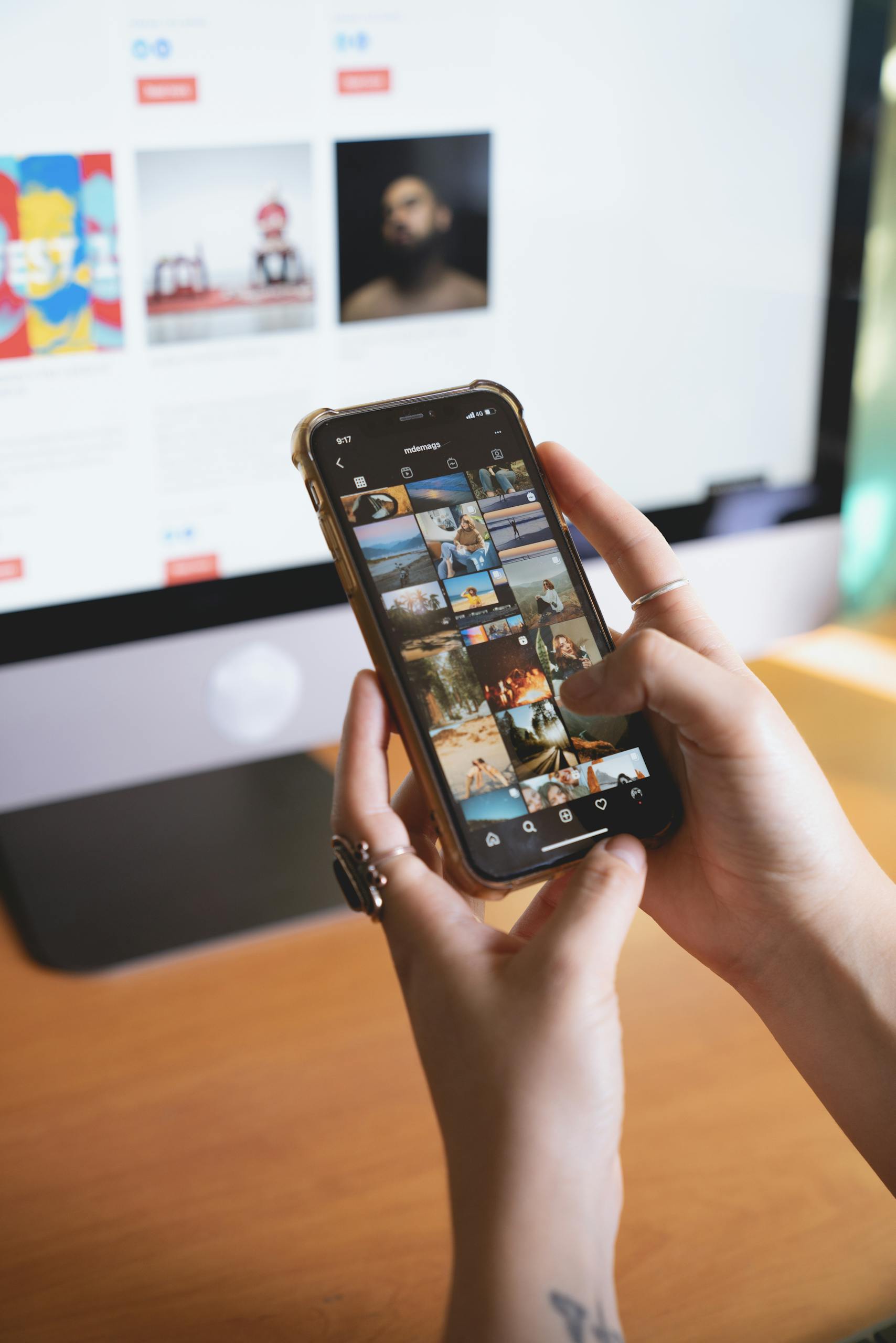 Close-up of a person browsing a smartphone with an Instagram feed, with a computer monitor in the background.